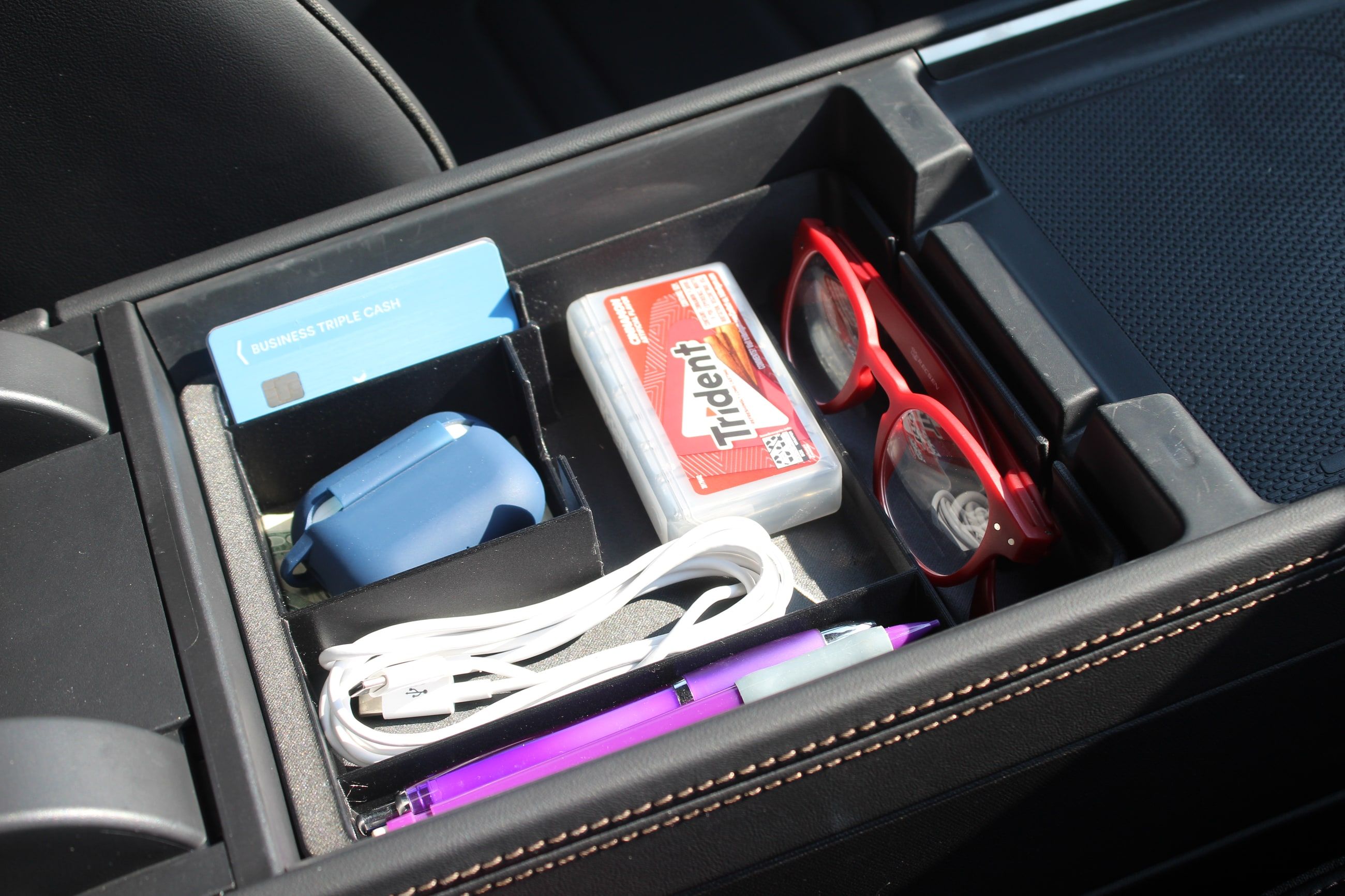 The Squared Away Fitted Tray seated in the Rivian R2 center console armrest cubby, loaded with a credit card, an earbud case, a pack of gum, red reading glasses, a coiled charge cord and a pen