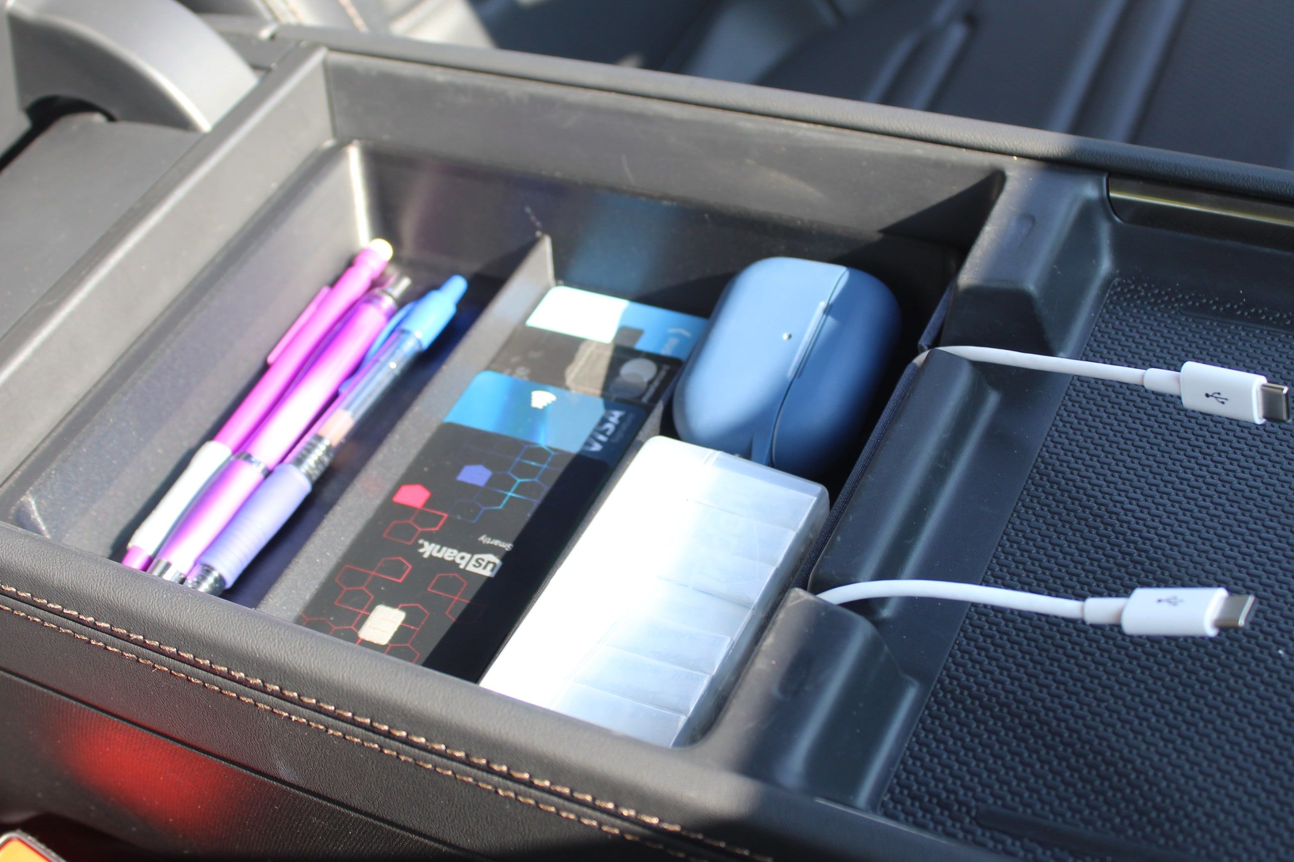 The Squared Away Tray · USB seated in the Rivian R2 center console armrest cubby, holding pens, cards, an earbud case and a pack of gum, with two white charge cords running out through the pass-through slots in the tray wall